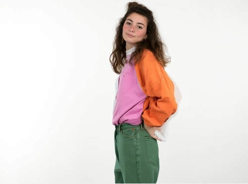 Smiling woman with wavy brown hair wearing a colorful pullover and green pants.